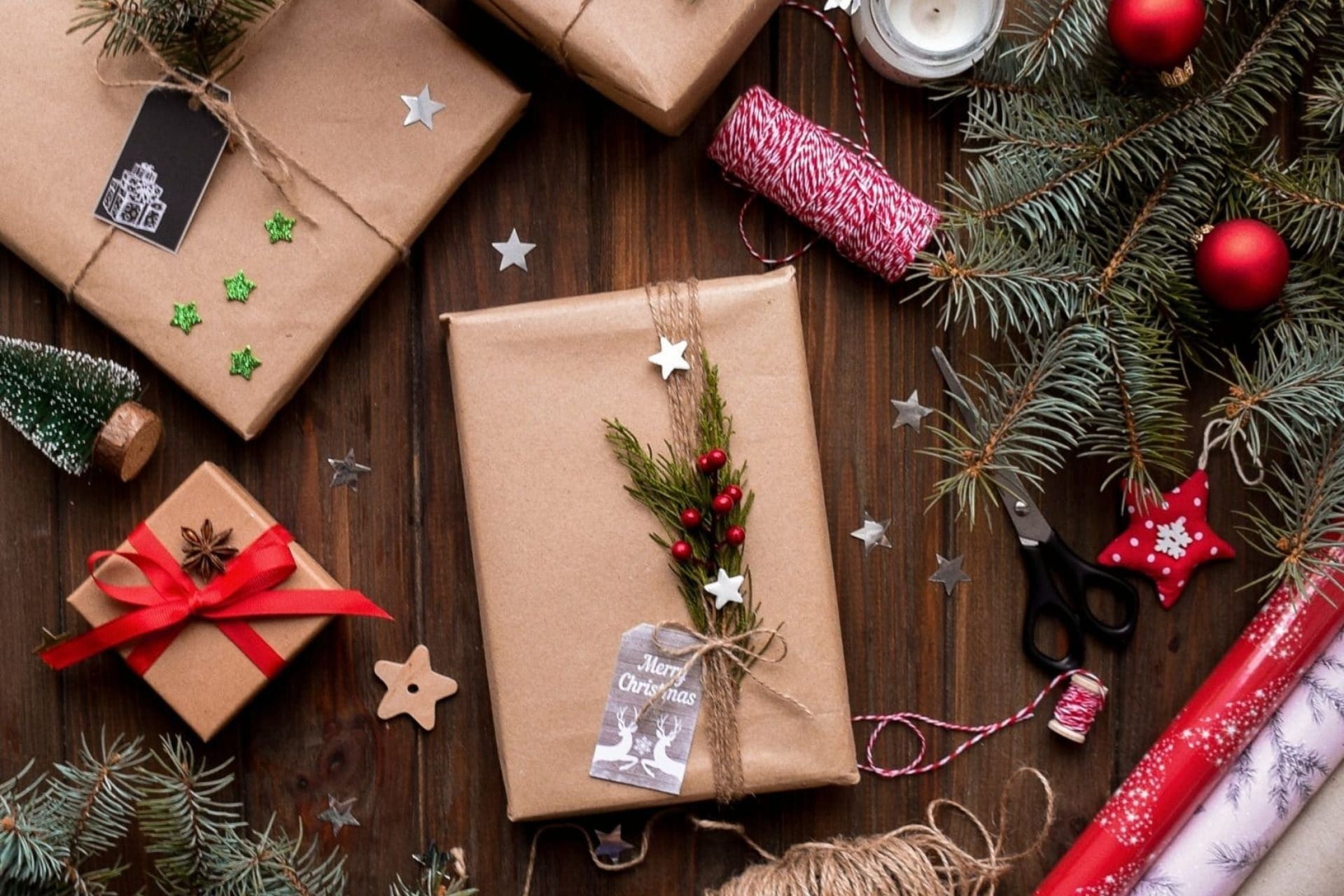 Mit Naturmaterialien verpackte Weihnachtsgeschenke, umgeben von Tannenzweigen, roten Kugeln, Geschenkpapier und Bastelmaterial – weihnachtlich, kreativ und nachhaltig.