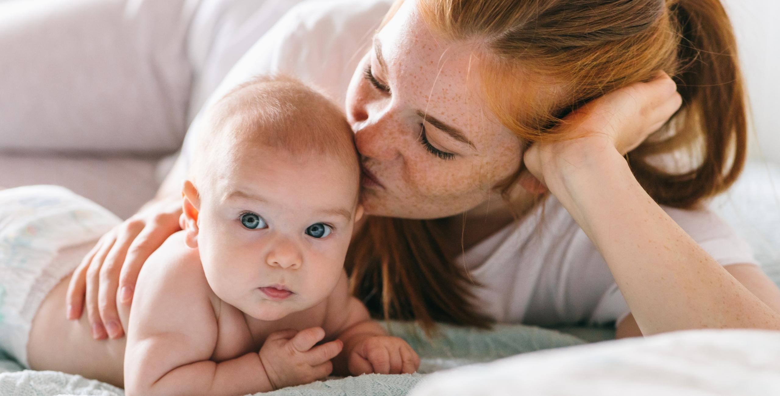 Liebende Mutter küsst ihr Baby beim Kuscheln auf dem Bett – inniger Moment voller Geborgenheit und Wärme
