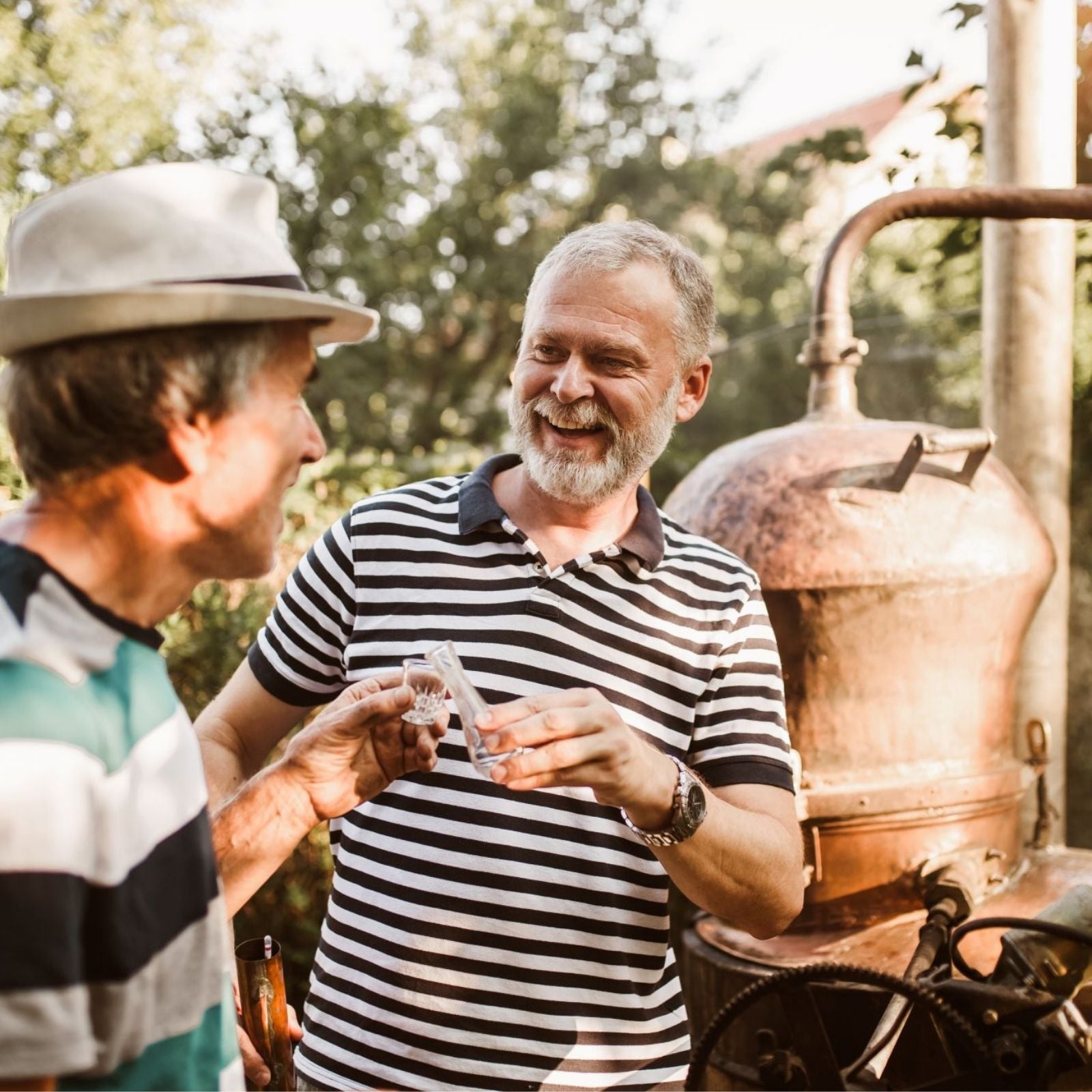 Zwei Männer an einer Kupferdestille lachen und probieren frisches ätherisches Öl – handwerkliche Herstellung in familiärer Atmosphäre