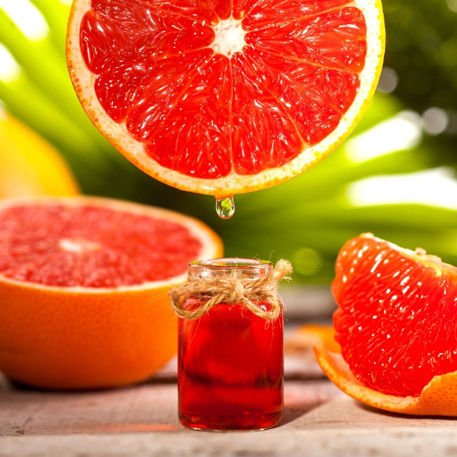 Aufgeschnittene rote Grapefruit mit Tropfen, der in ein kleines Fläschchen ätherisches Grapefruitöl fällt – Zitrusfrucht im Vordergrund, grüne Natur im Hintergrund