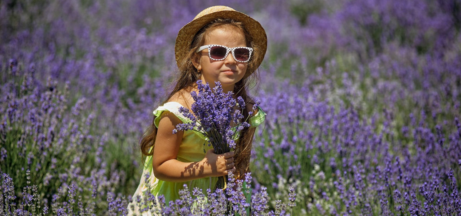 Ein fröhliches Mädchen mit Sonnenhut und Sonnenbrille steht lachend in einem Lavendelfeld und hält einen duftenden Strauß in der Hand.