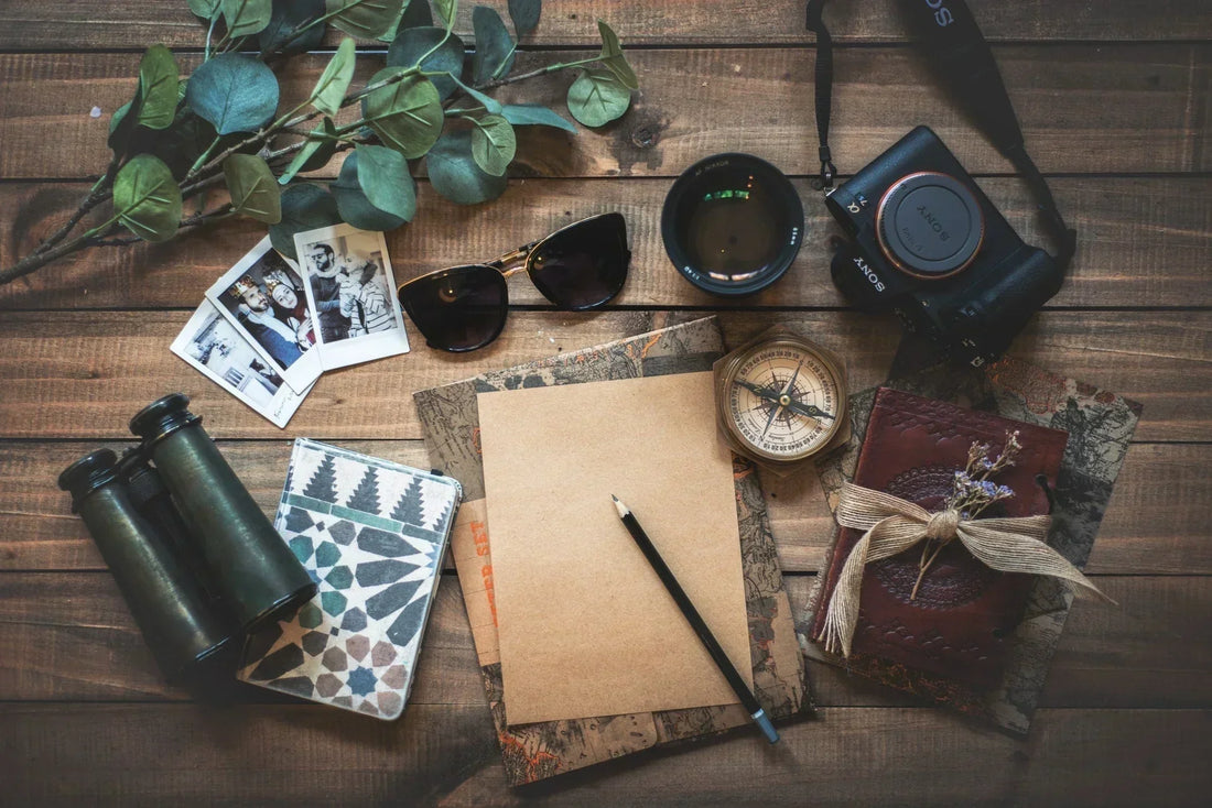 Reiseutensilien auf einem Holztisch – darunter Kamera, Kompass, Notizbuch, Sonnenbrille, Fotos, Fernglas und Lavendelzweig – als Symbol für Abenteuerlust, Planung und Naturverbundenheit.