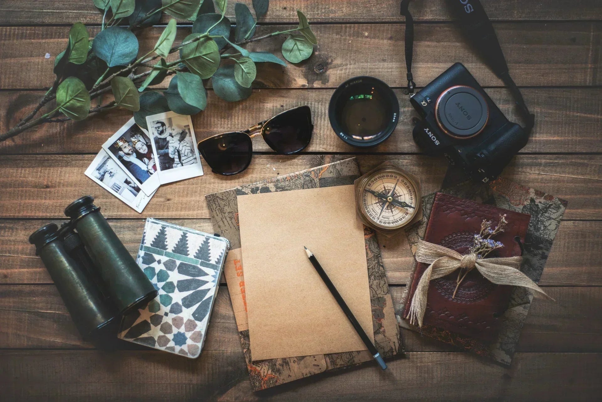Reiseutensilien auf einem Holztisch – darunter Kamera, Kompass, Notizbuch, Sonnenbrille, Fotos, Fernglas und Lavendelzweig – als Symbol für Abenteuerlust, Planung und Naturverbundenheit.