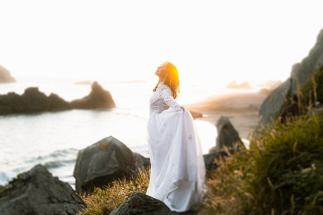 Eine Frau in einem weißen Kleid genießt die frische Brise an einer wilden Küste bei Sonnenuntergang. Ein Moment purer Freiheit, Naturverbundenheit und magischer Kraft – die Essenz von BREEZE einer ESSENCE-ORIGINALS Duftmischung.