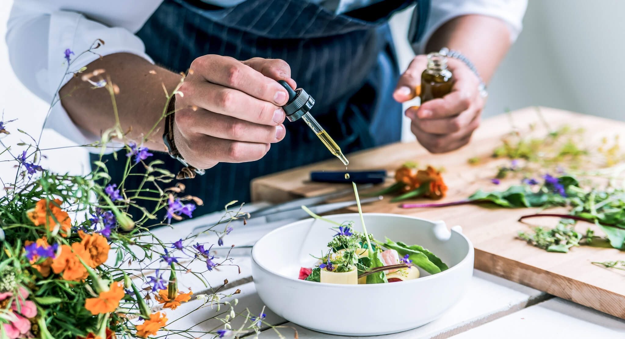 Ein Koch verfeinert ein kunstvoll angerichtetes Gericht mit ätherischem Öl aus der Pipette – aromatische Küche mit essbaren Blüten, Kräutern und viel Liebe zum Detail.