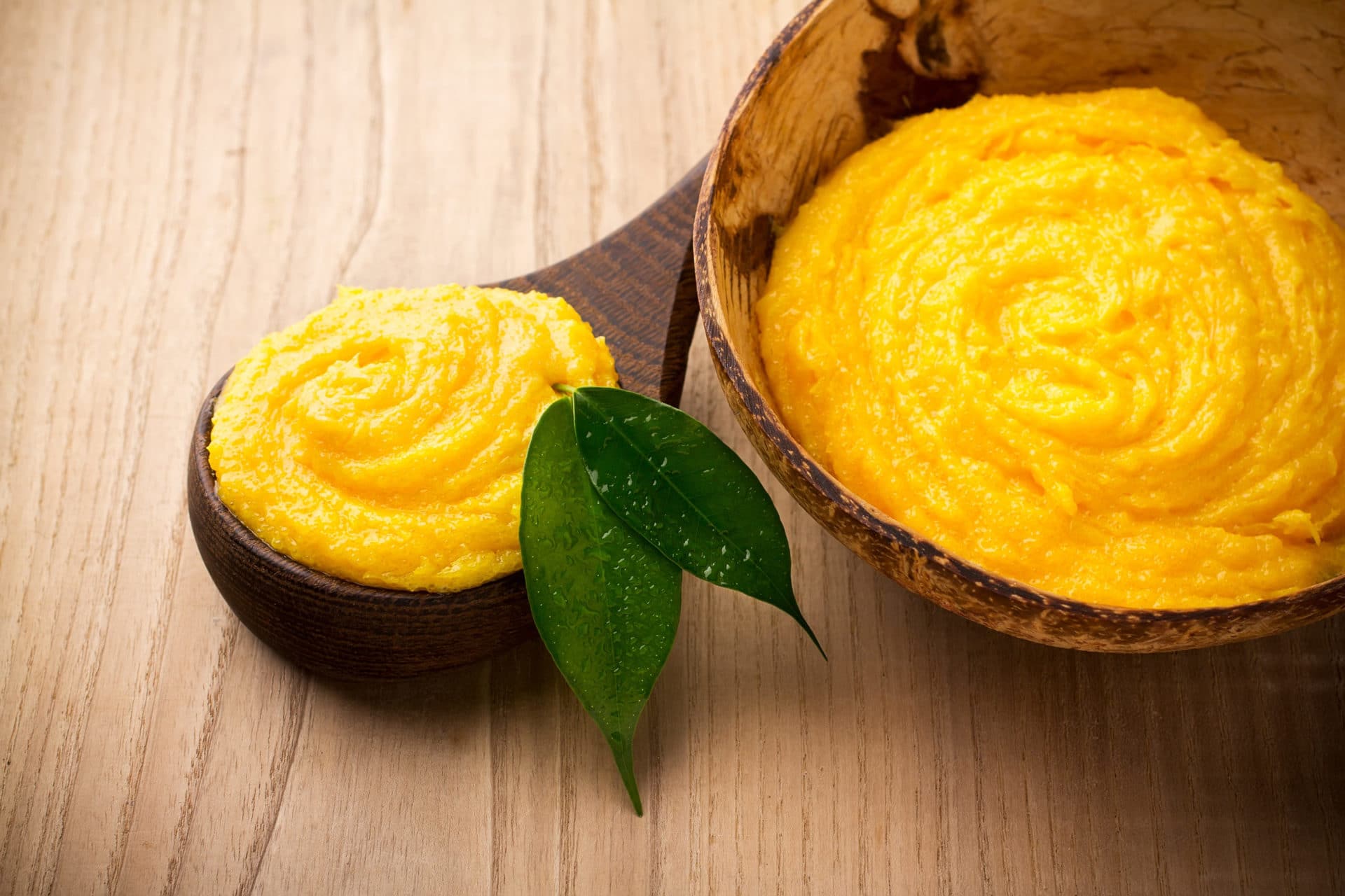 Cremige, goldgelbe Bio-Mangobutter in einer Kokosnussschale auf Holzuntergrund. Natürliche Feuchtigkeitspflege für Haut, Lippen und Haare.