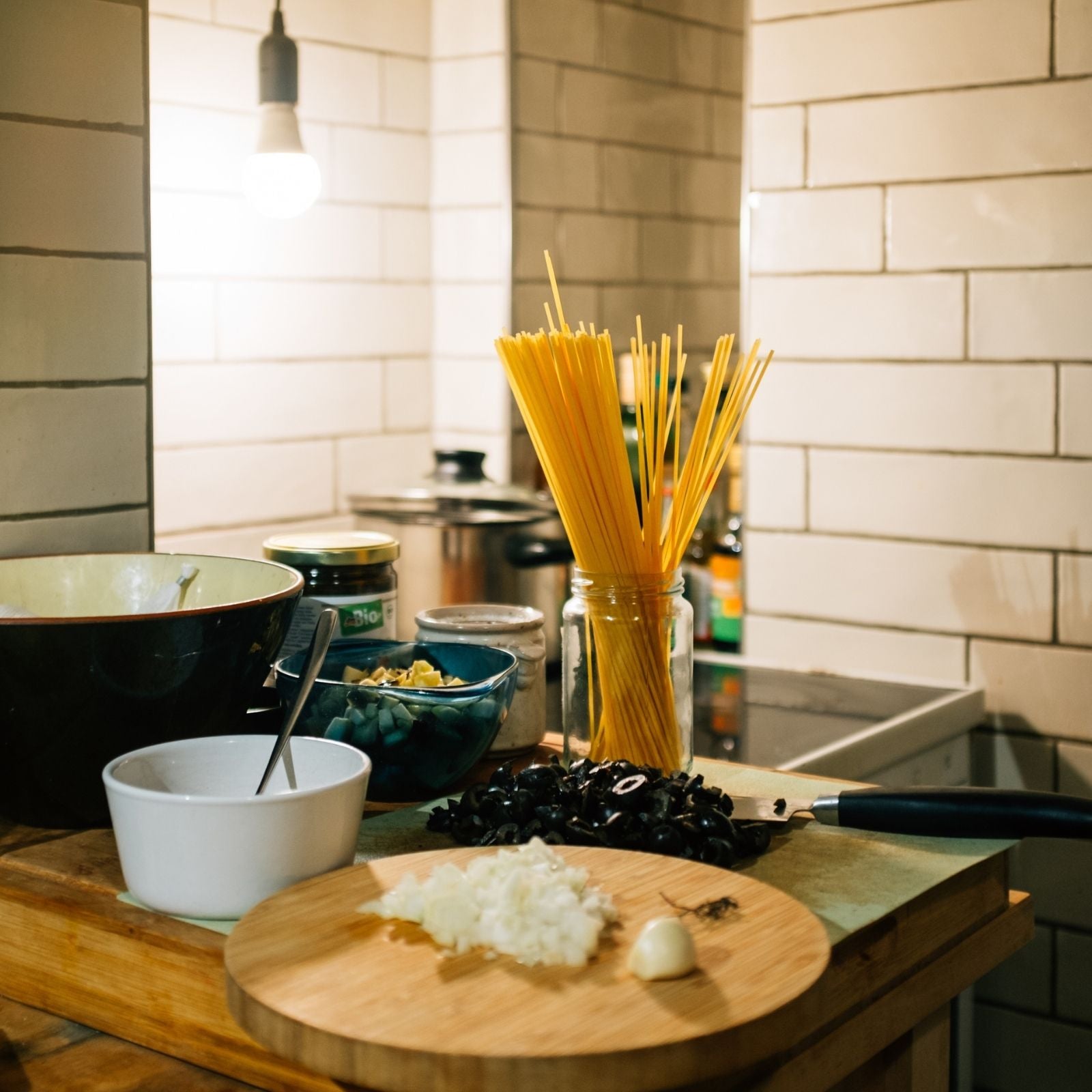 Ein stimmungsvoller Blick in eine gemütliche Küche mit Holzarbeitsfläche, Kochutensilien, Gewürzen und frischen Zutaten wie Pasta, Oliven und Zwiebeln – bereit für ein aromatisches Kochvergnügen mit ätherischen Ölen.