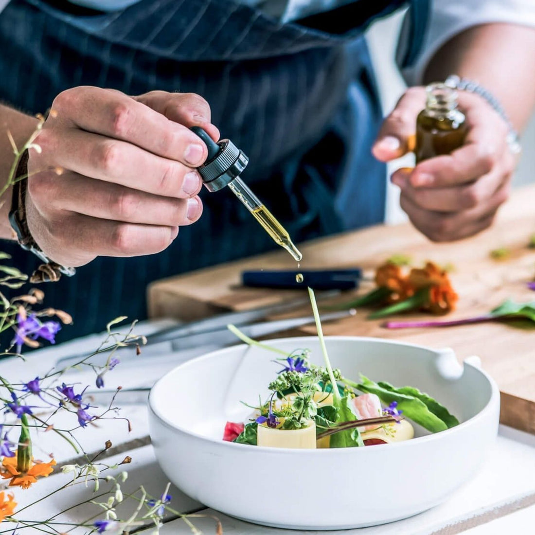 Ein Koch träufelt ätherisches Öl aus einer Pipette über ein kunstvoll angerichtetes Gericht mit essbaren Blüten – Aromaküche mit Pflanzenkraft