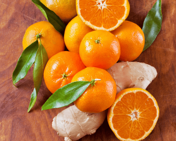 Frische Mandarinen mit grünen Blättern und aufgeschnittener Frucht neben Ingwer auf einem Holztisch – Symbol für die aromatische Vielfalt von ätherischem Mandarinenöl.