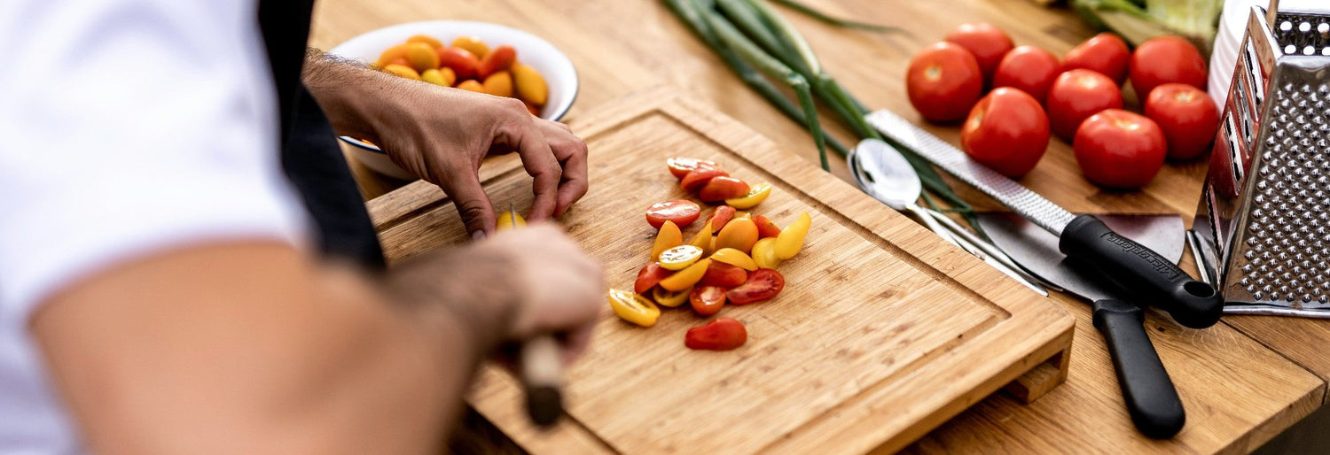 Frische Zutaten auf einem Holzbrett: Ein Koch schneidet bunte Kirschtomaten, daneben liegen Frühlingszwiebeln, Küchenutensilien und reife Tomaten – natürliche Aromaküche mit frischen Zutaten.
